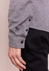 Nærbillede af en persons hånd, der hviler ved siden af kroppen, iført en grå tekstureret langærmet skjorte med sorte knapper og sorte bukser.