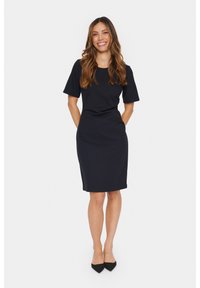 Robe ajustée bleu marine à manches courtes, col rond, longueur au genou et texture lisse. Associée à des chaussures noires à bout pointu.