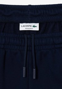 Detailní záběr na pas elastických tepláků tmavě modré barvy s nastavitelnými tkaničkami a bílou etiketou Lacoste, která zobrazuje zeleného krokodýla a nápis "Regular Fit".