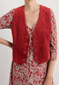 Gilet en velours côtelé rouge avec un col en V, comportant cinq boutons et une coupe courte. Porté par-dessus une robe à motifs floraux avec des accents blancs.
