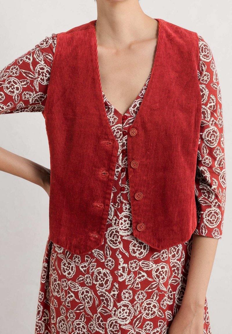 Gilet en velours côtelé rouge avec un col en V, comportant cinq boutons et une coupe courte. Porté par-dessus une robe à motifs floraux avec des accents blancs.