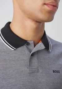 Pilkas polo marškinėliai su juodu apykakle, su baltais dryžiais, dviem mygtukais ir mažu "BOSS" logotipu ant krūtinės. Švelni tekstūra.