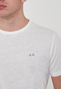 T-shirt bianco a maniche corte con collo rotondo, caratterizzato da una consistenza morbida e un piccolo logo geometrico blu sul lato sinistro del petto.