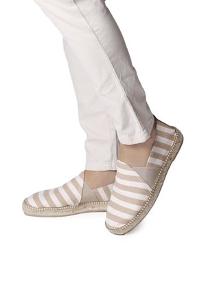 Espadrilles a rayas con parte superior de lona beige y blanca, paneles laterales elásticos y suela de cuerda de yute. Llevadas con pantalones blancos.