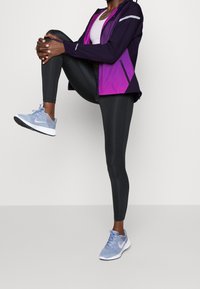 Chaqueta deportiva morada y negra con acentos reflectantes, combinada con mallas negras y zapatillas de deporte azul claro con un logo rosa.