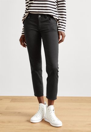 Persoon draagt zwarte slim-fit cropped broek, een zwart-wit gestreept shirt met lange mouwen en witte hoge sneakers, staand op een houten vloer.