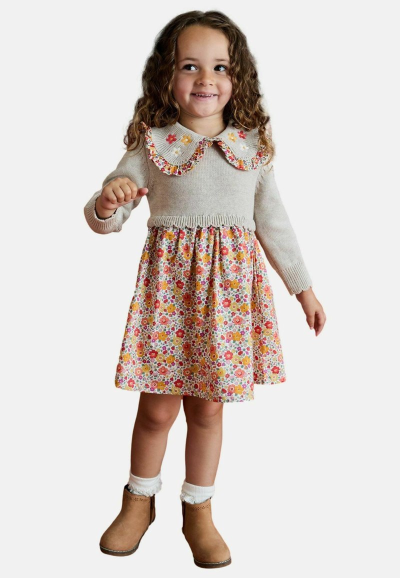 Lächelndes junges Mädchen mit lockigem Haar, das einen beigen Pullover mit floralem Kragen über einem passenden Blumenkleid und beigen Stiefeletten trägt.