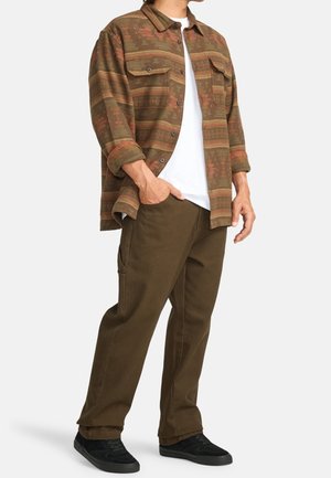 Mand iført brun mønstret skjorte med knapper over hvid t-shirt, brune bukser med sidelomme og sorte sko, stående med den ene hånd i lommen.