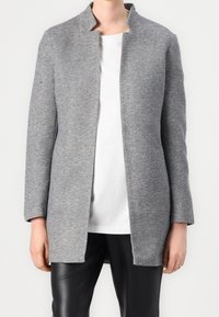 Cappotto grigio in misto lana con colletto alto, maniche lunghe e design minimalista, indossato sopra una maglietta bianca e pantaloni in pelle nera.