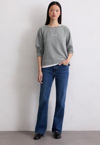 Sudadera gris con puños acanalados y cuello redondo, que presenta un pequeño logo. Combinada con jeans azules de campana y zapatos negros, de pie contra una pared clara.
