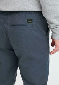 Marineblå bukser laget av teksturert stoff, med en baklomme og en liten svart merkelapp sydd fast på midjebåndet.