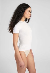 Maglietta in cotone bianco aderente con maniche corte, scollo rotondo e texture liscia, indossata con biancheria intima color nudo, mostrata di profilo.