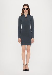 Rochie gri cu mâneci lungi, cu guler cu fermoar, design ajustat și textură netedă. Asortată cu pantofi cu vârf ascuțit negri și ochelari de soare.