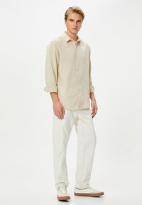 Beige linnen overhemd met knoopsluiting en opgerolde mouwen, gecombineerd met witte broek en lichtgekleurde sneakers. Eenvoudig ontwerp met een zachte textuur.