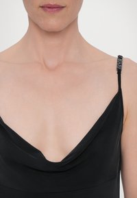 Rochie neagră cu decolteu tip cowl și curele subțiri, ajustabile, însoțite de accesorii decorative, evidențiind o textură de țesătură netedă și moale.