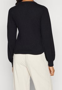 Pull noir en maille avec manches longues, poignets et ourlet côtelés, présentant une coupe ajustée et un léger détail aux épaules. Design classique et polyvalent.