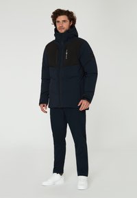 Marinblå puffjacka med en svart övre panel, med dragkedjefickor och hög krage, som bärs med matchande marinblå byxor och vita sneakers.