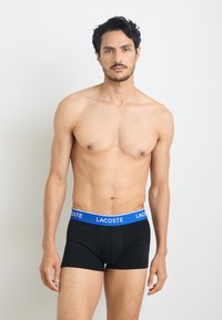 Black boxer shorts with a blue Lacoste logo waistband, featuring a fitted design and soft material, showcased on a model.