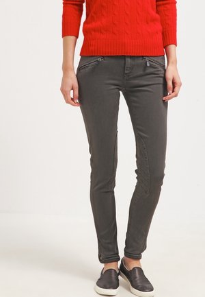 Osoba nosící tmavě šedé štíhlé kalhoty s kapsami na zip vpředu, červený svetr s pleteným vzorem a černé nazouvací boty s bílými podešvemi.