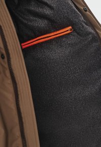 Brązowa kurtka z teksturowanym ciemnym podszewką, z pomarańczową kieszenią na zamek błyskawiczny wewnątrz. Tkanina wydaje się gładka i wytrzymała.