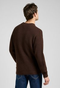 Brun stickad tröja med ribbat mönster, rund halsringning och avslappnad passform. Tydligt sänkt axlar och en subtil fåll.