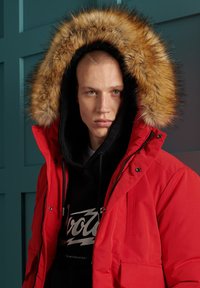 Rød isoleret parka med aftagelig pelsforet hætte, der har lynlås foran, store lommer og sort indvendig hætte med grafisk design.