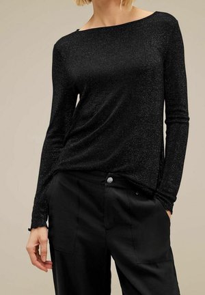 Vrouw draagt een nauwsluitend zwart topje met lange mouwen met subtiele glans en zwarte high-waisted broek met voorzakken tegen een beige achtergrond.