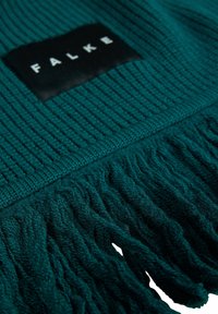 Sciarpa in maglia color teal caratterizzata da un motivo a coste testurizzato, dettaglio frangiato sul fondo e un'etichetta nera con il marchio "FALKE" impresso in bianco.