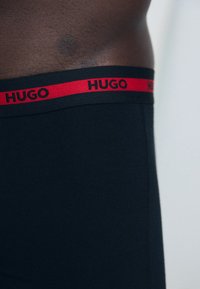 Schwarze Boxer-Shorts mit einem roten elastischen Bund, auf dem "HUGO" steht. Der Stoff wirkt glatt und das Design ist körperbetont.