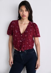 Červený kvetinový crop top s krátkymi puff rukávmi, výstrihom do V a detailom s uzlom vpredu, spárovaný s tmavými vysokými džínami.