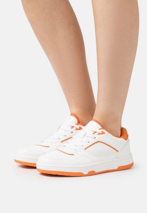 Baskets blanches avec des accents orange portées pieds nus, présentant des vues de côté et de face sur un fond blanc uni.