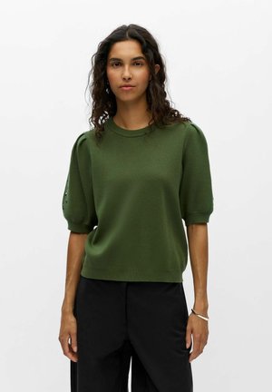 Femme aux cheveux bouclés et foncés portant un pull vert à manches courtes et un pantalon noir, debout devant un fond blanc uni.