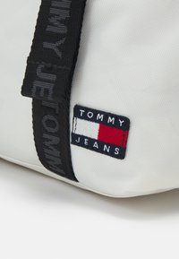 Gros plan d'un sac en tissu blanc avec une sangle noire et un patch logo Tommy Jeans rectangulaire en rouge, blanc et bleu marine.