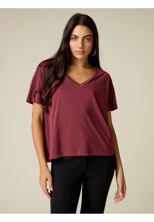 Femme aux longs cheveux foncés, portant des boucles d'oreilles créoles dorées, un haut ample bordeaux à col en V et manches courtes, et un pantalon noir sur un fond uni.