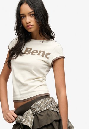 Ung kvinde med langt mørkt hår iført en hvid Bench T-shirt og en ternet skjorte bundet omkring taljen.