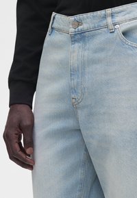 Main à la peau foncée reposant à côté d'un jean en denim bleu clair avec bouton et fermeture éclair, portée avec un haut noir à manches longues.