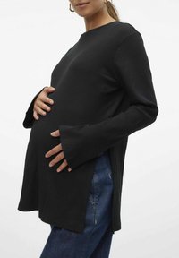 Svart ribbat graviditetstopp med långa, utsvängda ärmar och sidoflikar. Matcher med blå denimbyxor som visar en åtsittande silhuett.