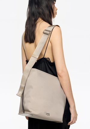 Beżowa torba na ramię wykonana z gładkiego materiału. Posiada szeroki pasek i zamknięcie na zamek u góry, z logo nadrukowanym na boku.