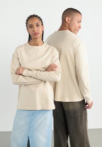 Beige long-sleeve t-shirt with crew neck, displayed on two models, paired with a light blue denim skirt and dark jeans, simple design.