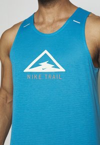 Homme portant un débardeur bleu Nike Trail avec un logo montagne blanc et un motif d'éclair sur la poitrine.