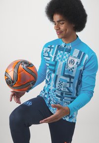 Maglietta sportiva blu con motivi geometrici, maniche lunghe e zip a metà, abbinata a pantaloni blu navy. Una vibrante palla da calcio arancione è bilanciata sulla sua mano.