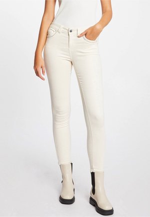 Femme portant un jean ajusté blanc cassé et des bottines beiges avec des panneaux élastiques noirs sur les côtés, sur fond blanc.