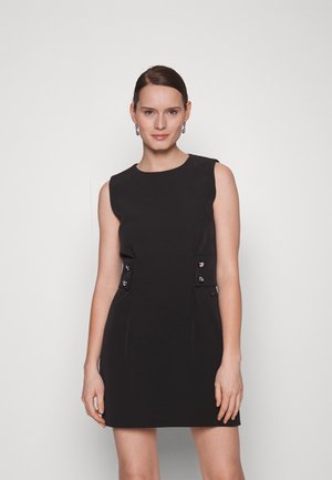 Γυναίκα με σκουλαρίκια, ντυμένη με ένα αμάνικο μαύρο φόρεμα με κουμπωμένες λεπτομέρειες στη μέση, που στέκεται μπροστά σε μια απλή φωτεινή στάθμη.