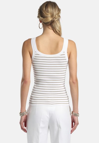 Femme aux cheveux blonds coiffés en queue de cheval basse torsadée, portant un haut blanc sans manches côtelé avec de fines rayures horizontales grises et un pantalon blanc, vue de dos.