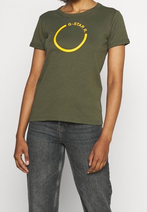 Osoba nosząca oliwkowo zielony t-shirt z żółtym pękniętym kołem i napisem „G-STAR”, w połączeniu z wyblakłymi czarnymi jeansami.