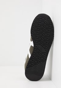 Buty sportowe z cholewką z siateczki w kolorze oliwkowej zieleni i bieli, czarną gumową podeszwą o fakturze z falistym wzorem oraz wzmocnionym noskiem dla zwiększenia trwałości.