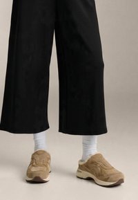 Beige ruskindsneakers med blødt for, en tyk hvid sål, kombineret med hvide ribstrikkede sokker og sorte bukser med vide ben.