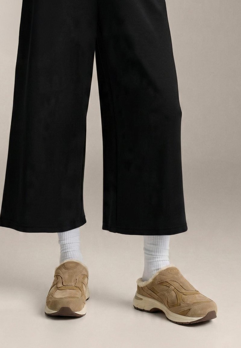 Beige ruskindsneakers med blødt for, en tyk hvid sål, kombineret med hvide ribstrikkede sokker og sorte bukser med vide ben.