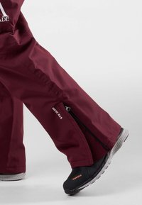 Pantaloni impermeabili color bordeaux con aperture laterali a zip, caratterizzati da un design elegante e un dettaglio logo. Indossati con calzature nere, evidenziando uno stile funzionale.