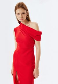 Vestido rojo de un solo hombro con tela drapeada, silueta ajustada y una apertura lateral. Textura suave y diseño simple sin patrones visibles.